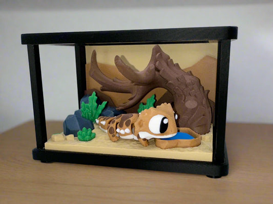 3D printed bearded dragon figure displayed inside a miniature terrarium with desert background, log hide, plants, and water dish.