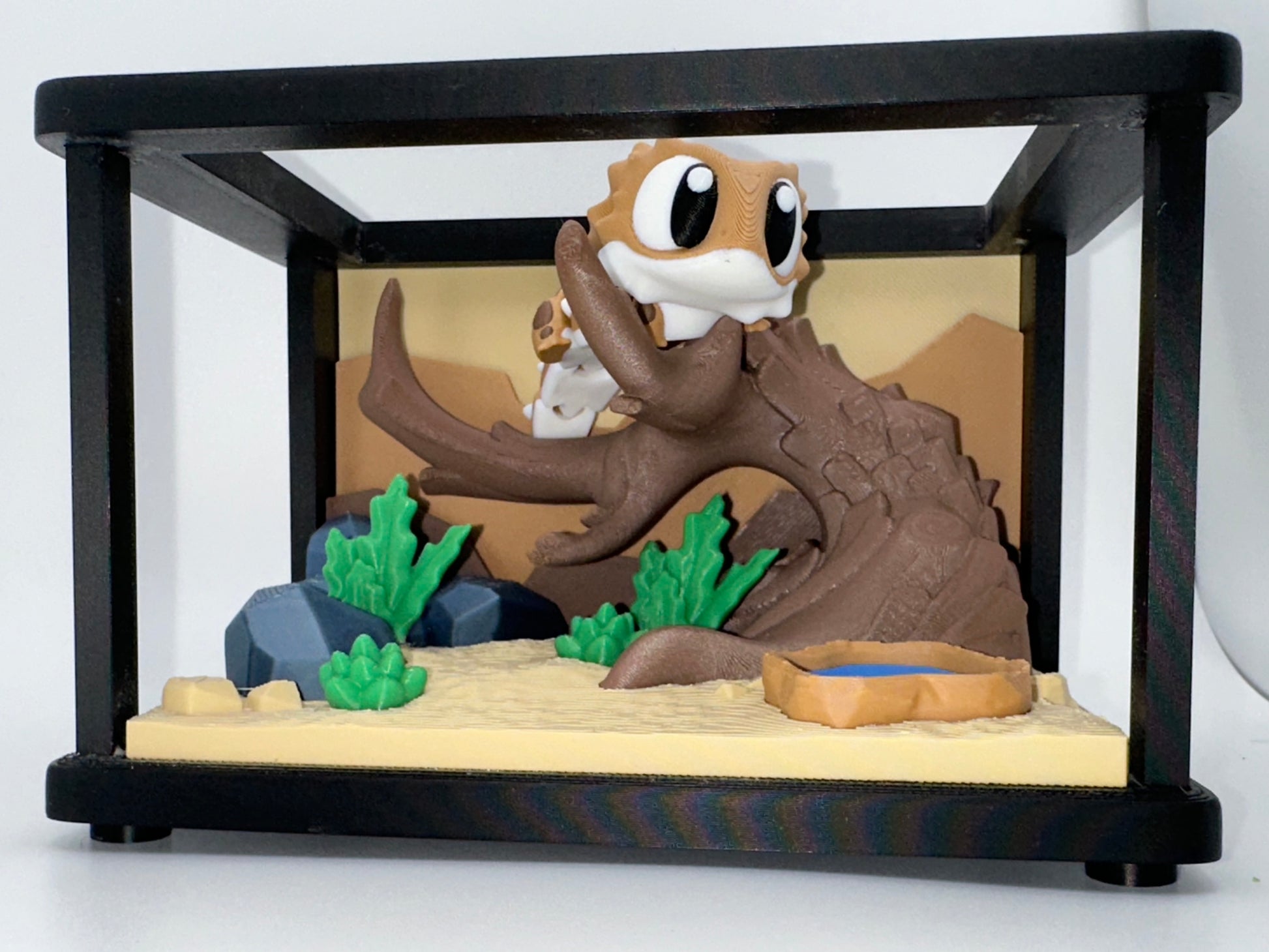3D printed bearded dragon figure displayed inside a miniature terrarium with desert background, log hide, plants, and water dish.