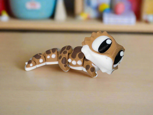 Close-up of articulated 3D printed bearded dragon figure with big eyes and brown-and-white patterned body on a tabletop.